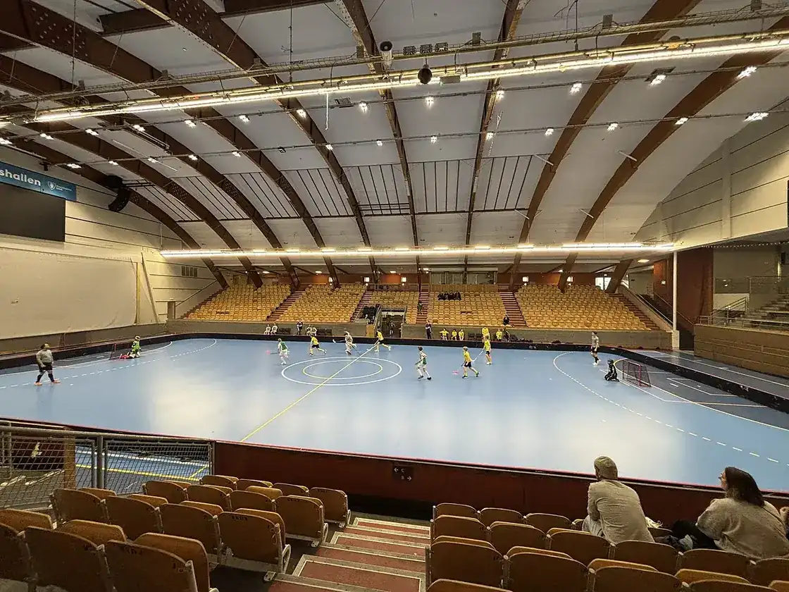 Sporthall med blå spelplan, spelare på golvet och tomma träläktare i förgrunden